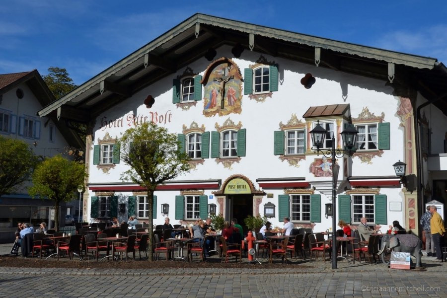 People at cafe tables sit in front of the Hotel Alte Post in Oberammergau, Germany on a sunny day. The sky is blue, and the building is painted with religious murals. The walls are white and the shutters are green.