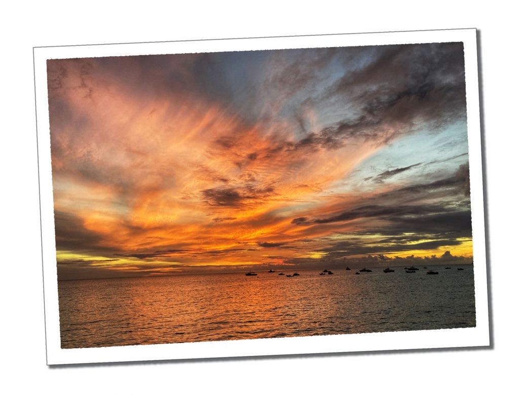 A fiery orange sky above tiny black silhouettes of boats on a calm evening sea