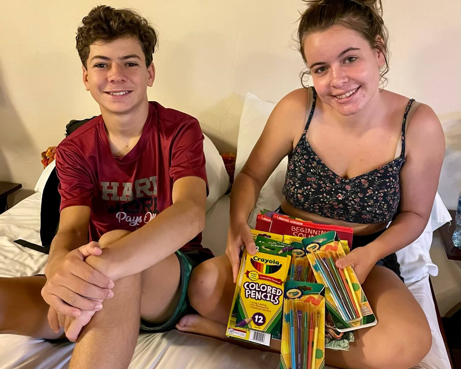 Boy and girl sitting on bed with girl displaying colored pencil, paintbrushes, books and other supplies