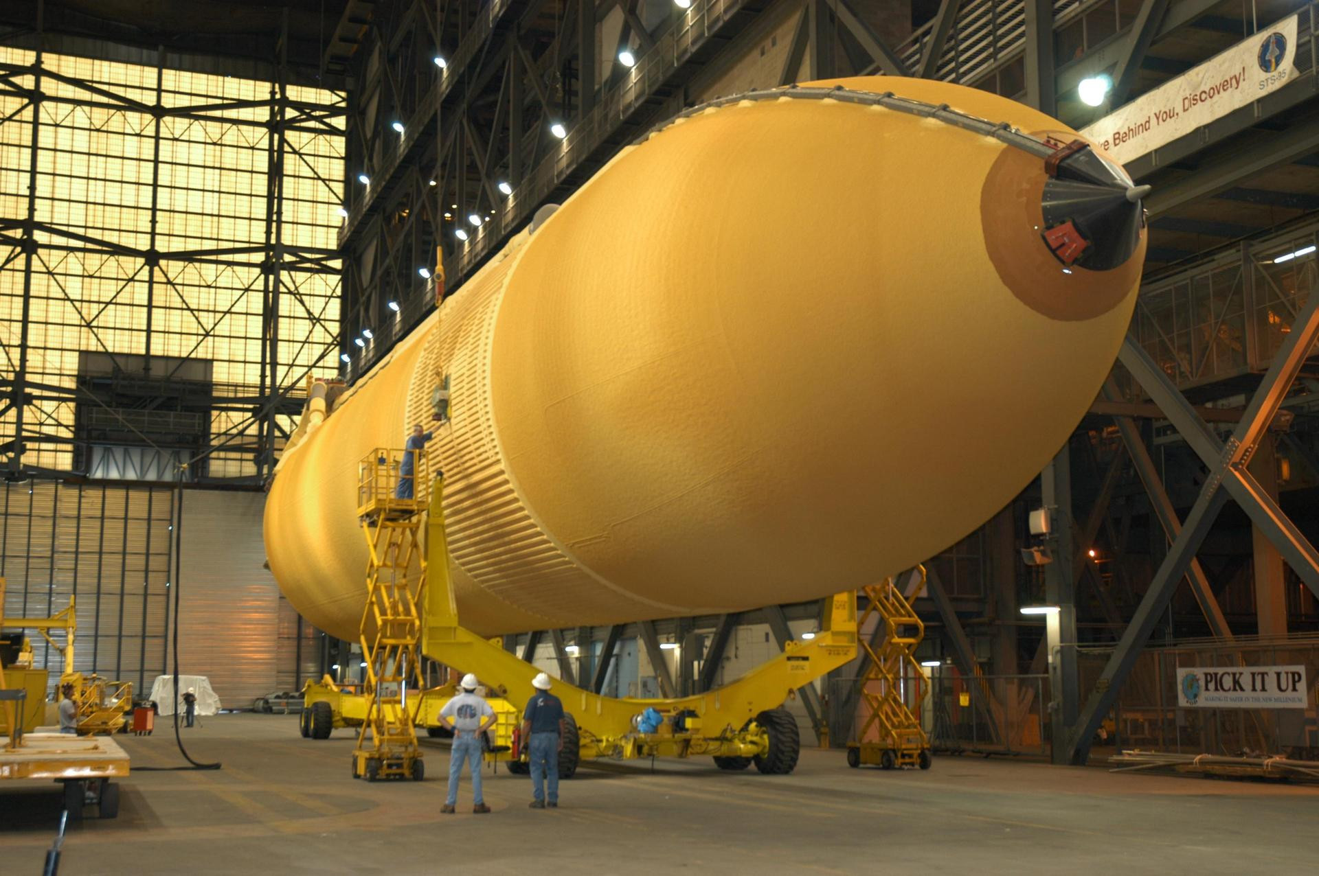 Workers in the transfer aisle of the Vehicle Assembly Building check the progress of External Tank 118 (ET-118) as it is lowered onto the transporter below it.