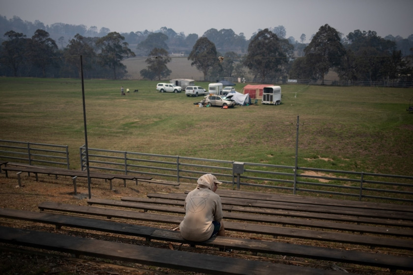 A man sitting on a bench among caravans and tents of evacuees in Cobargo, Australia, highlighting the mental and emotional toll of the bushfires on affected communities and travelers
