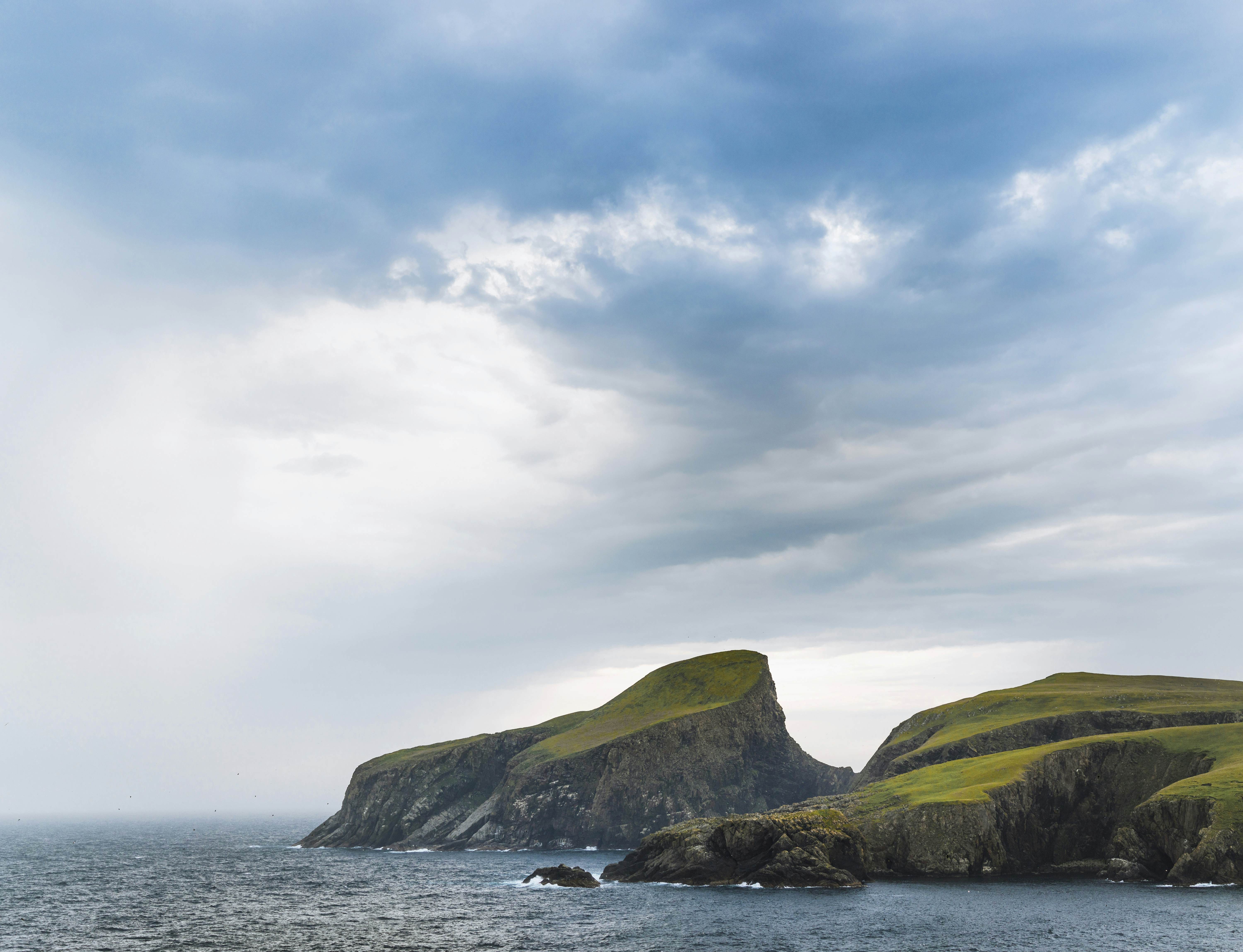 Green coastal cliffs set in dark, choppy waters, with cloudy gray skies overhead. 
