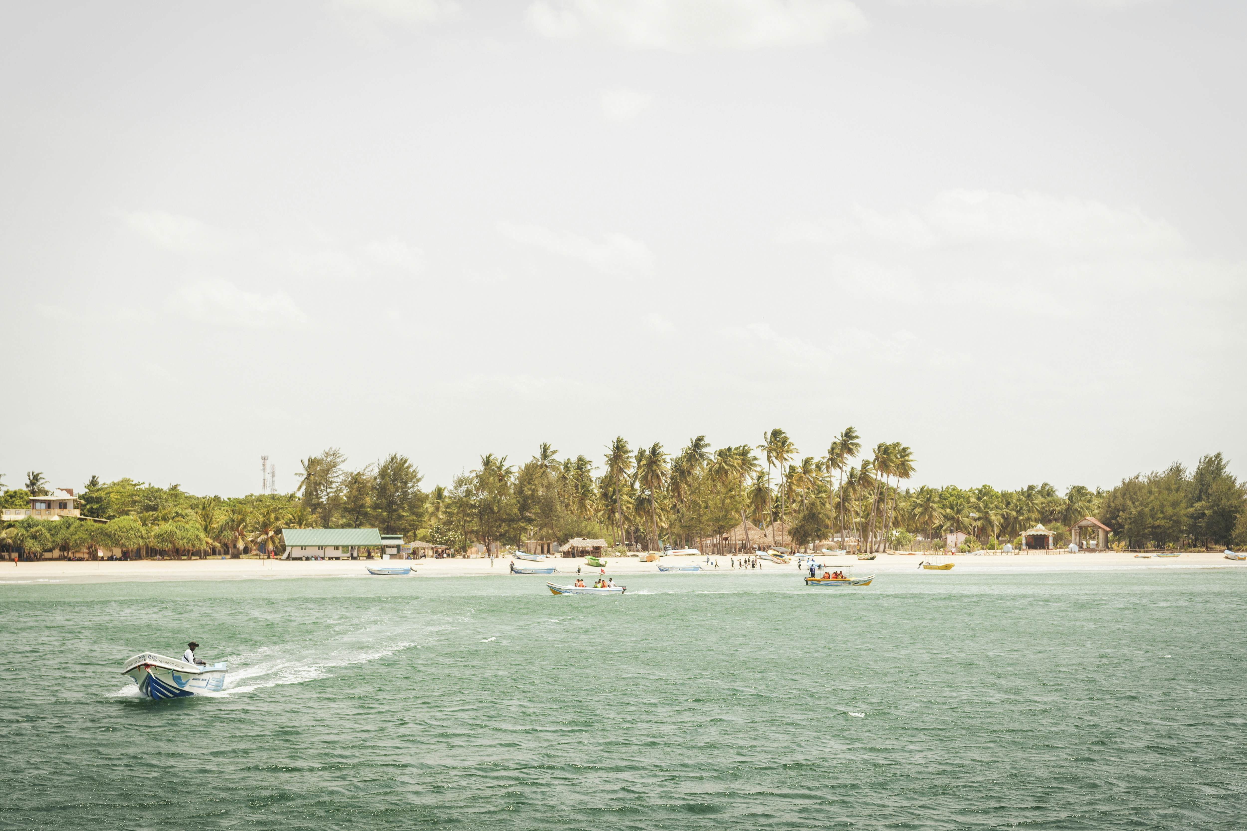 Boats in pale blue water leaving a beach on the shore with sparse greenery.
