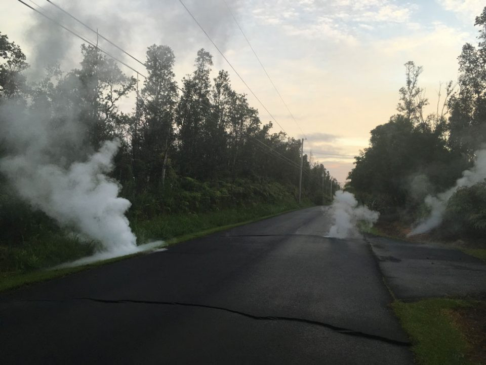 A road winds through a verdant landscape, with white gas venting from road cracks near Kilauea volcano in Hawaii.