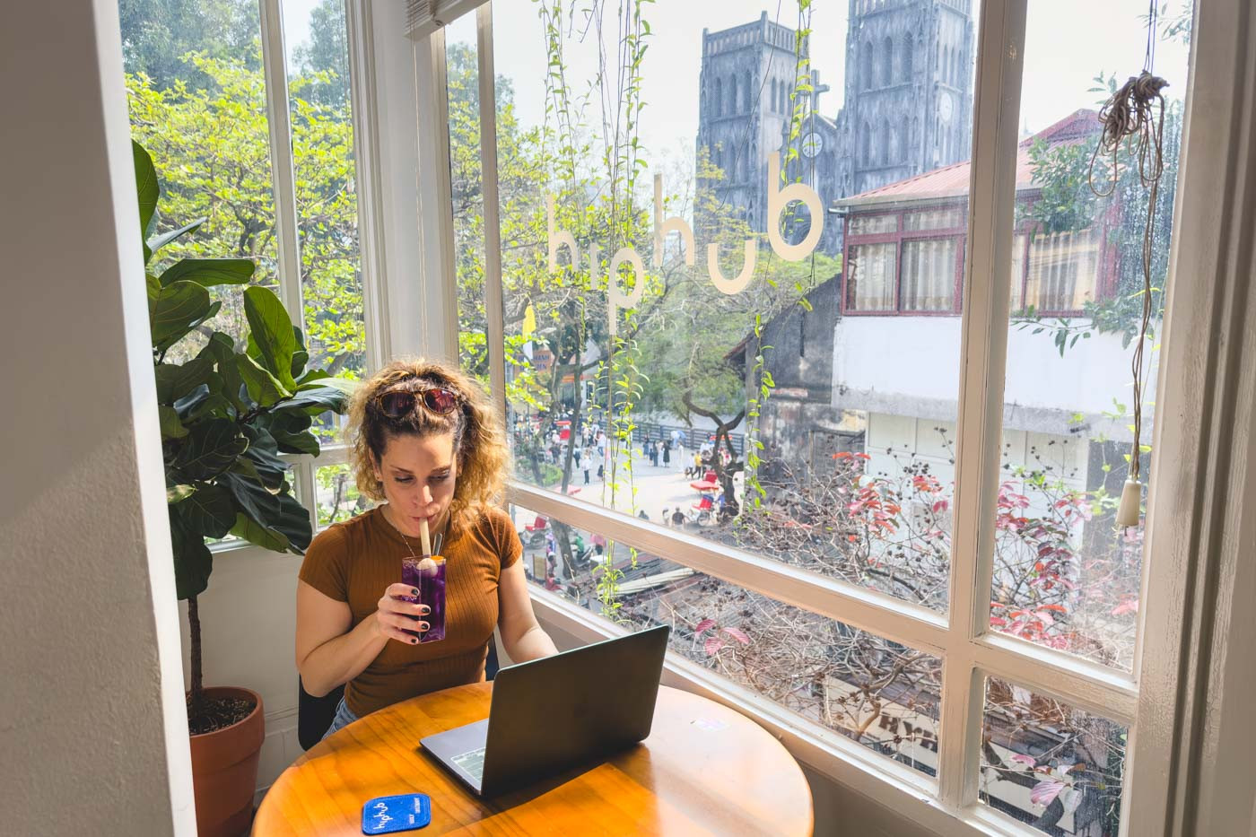 Nina sitting on a laptop and sipping on a purple drink inside HipHub Cafe with a view over Hanoi
