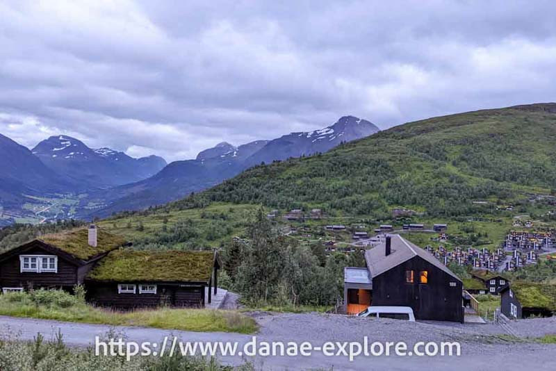 Dark wooden houses, with landscaped roofs, in a valley with green vegetation and mountains with a little snow in the background. Norway, is it expensive to travel in Norway?