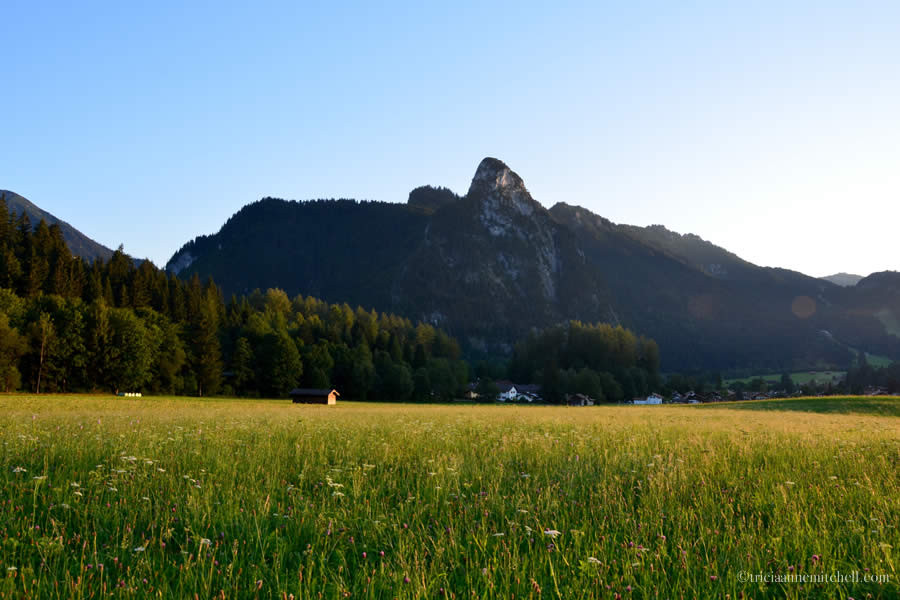 A pointed mountain towers over a yellow and green meadow in Oberammergau, Germany. You can see a small barn off in the distance.