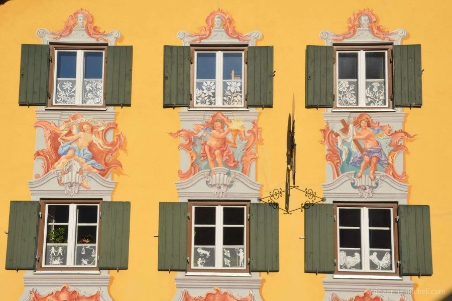 Close-up of a traditional Bavarian home: The wall is painted yellow, there are 6 green shutters, and murals of angels.