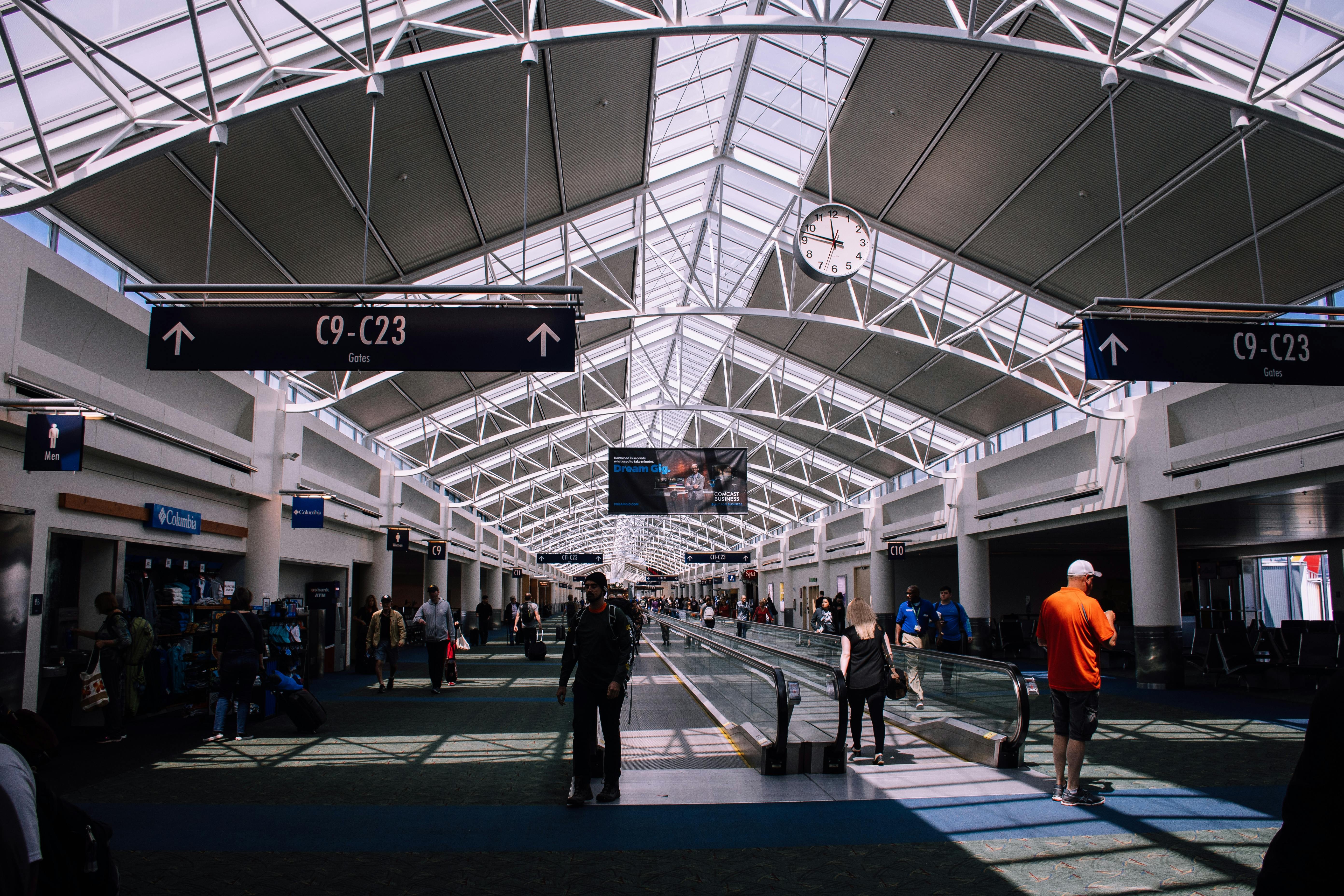 A busy airport terminal with passengers walking under a glass ceiling, large gate signs, a clock, and a moving walkway.