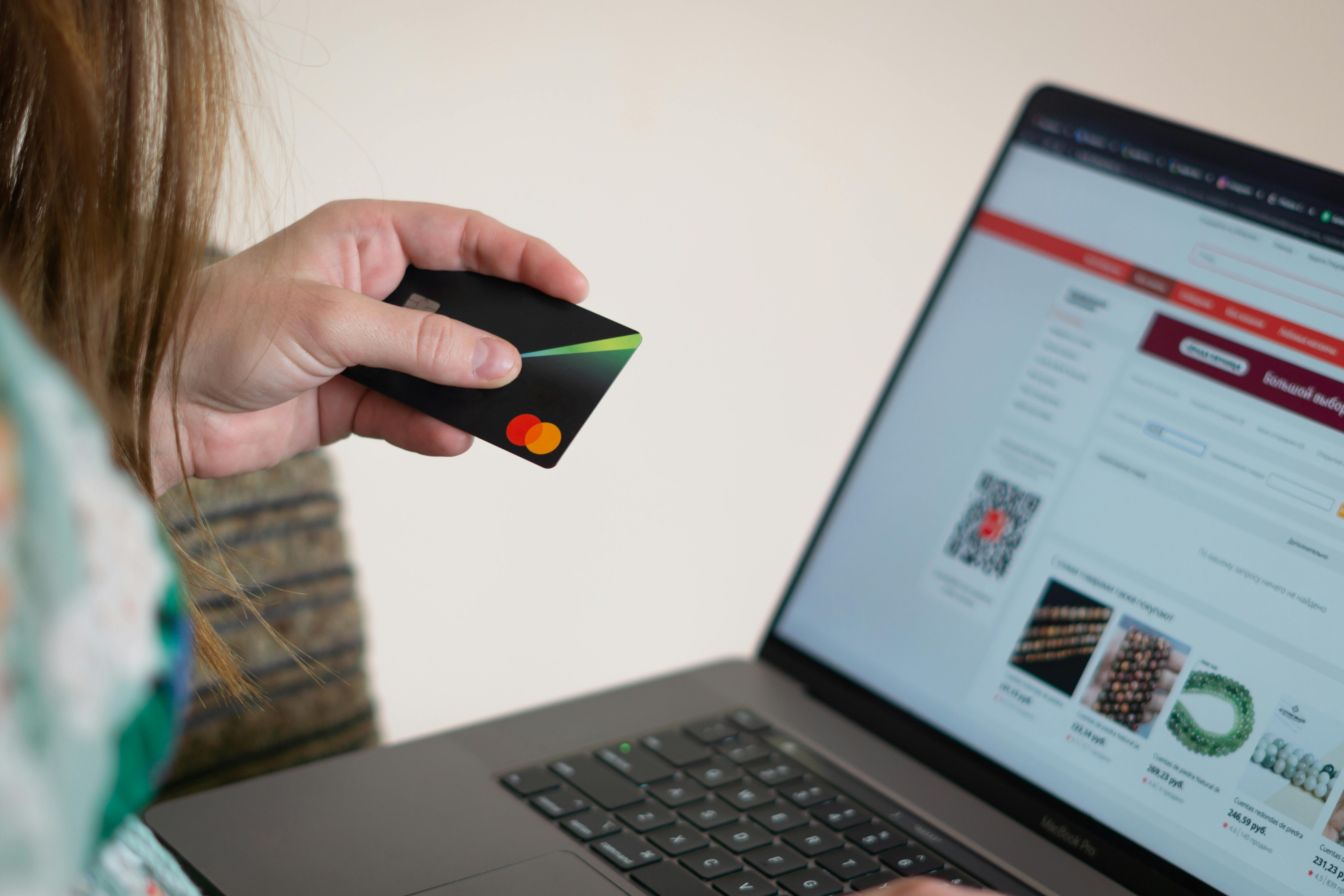 A person holding a credit card while shopping online on a laptop, with an e-commerce website displaying various products on the screen, showcasing the convenience of online transactions for travel planning.