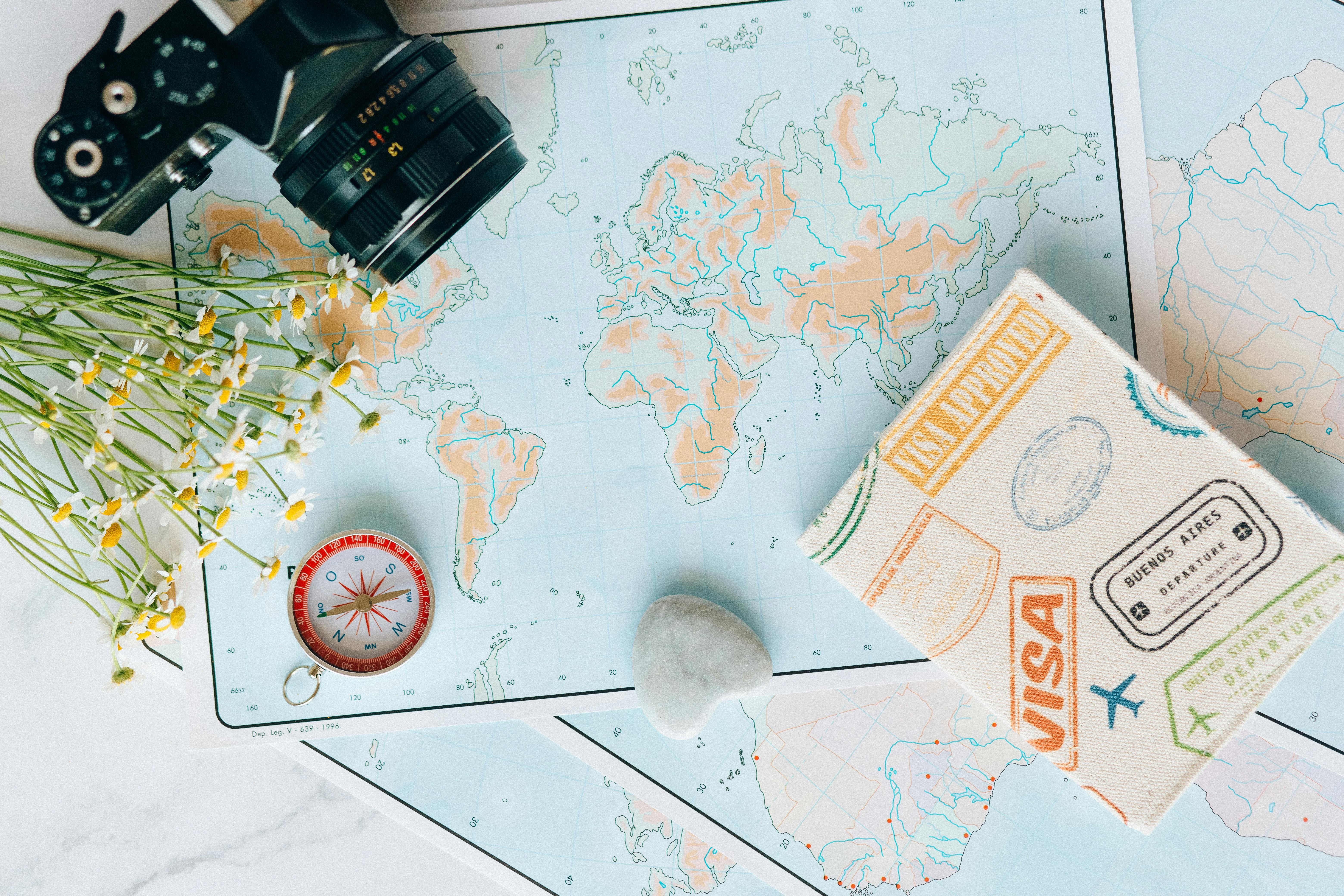 A travel-themed flat lay with a world map, vintage camera, compass, flowers, and a passport holder decorated with colorful stamps.