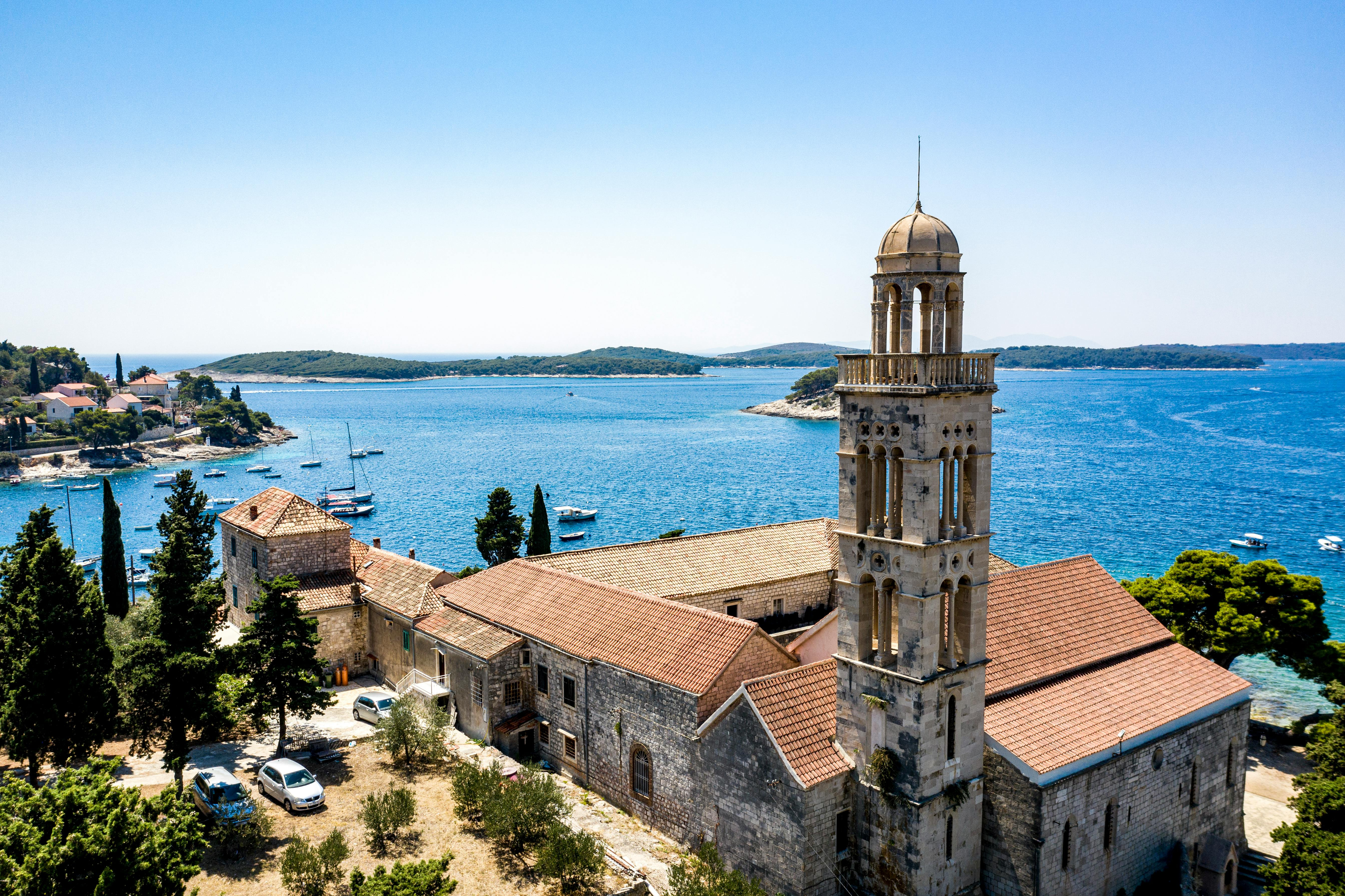 Historic stone church with a tall bell tower overlooking the blue sea, surrounded by trees, boats, and small islands in the background.