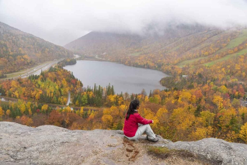 A person in a bright red jacket sits on a rocky ledge overlooking a scenic valley in autumn, one of the best places to visit in the fall, offering breathtaking views of colorful foliage and serene lakes