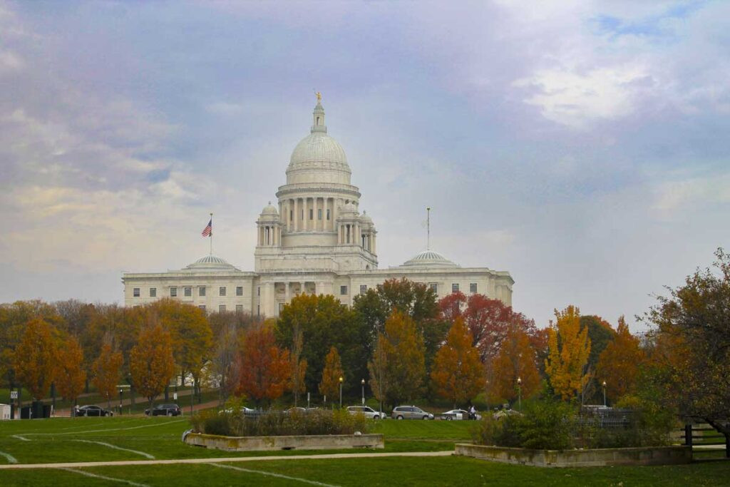 A majestic white-domed government building surrounded by trees in autumn colors stands against a cloudy sky. Cars are parked in front, and well-kept lawns and pathways are visible in the foreground—truly one of the best places to visit in the fall, showcasing vibrant colors and historical architecture