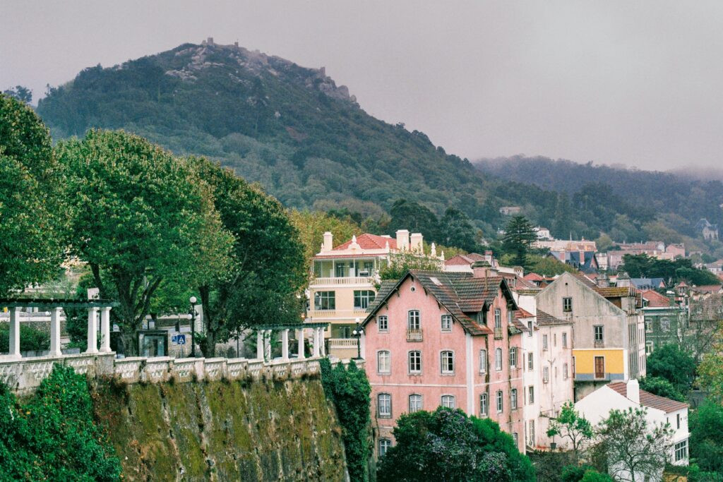 Colorful pastel houses in Sintra, Portugal with a misty forested mountain and ancient castle ruins in the background, framed by lush green trees and a moss-covered stone wall.