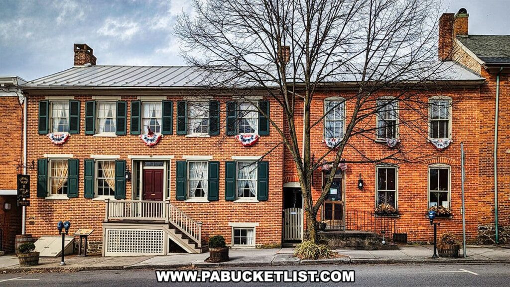 The front facade of the Shriver House Museum in Gettysburg, Pennsylvania, is captured in this image. It features a two-story red brick building with green shutters and a metal roof. Each window on the second floor is adorned with patriotic bunting, and a leafless tree stands in front, suggesting the photo was taken in a cooler season. The entrance has a red door accessible by a set of steps with a ramp, and a small sign beside the door indicates the building