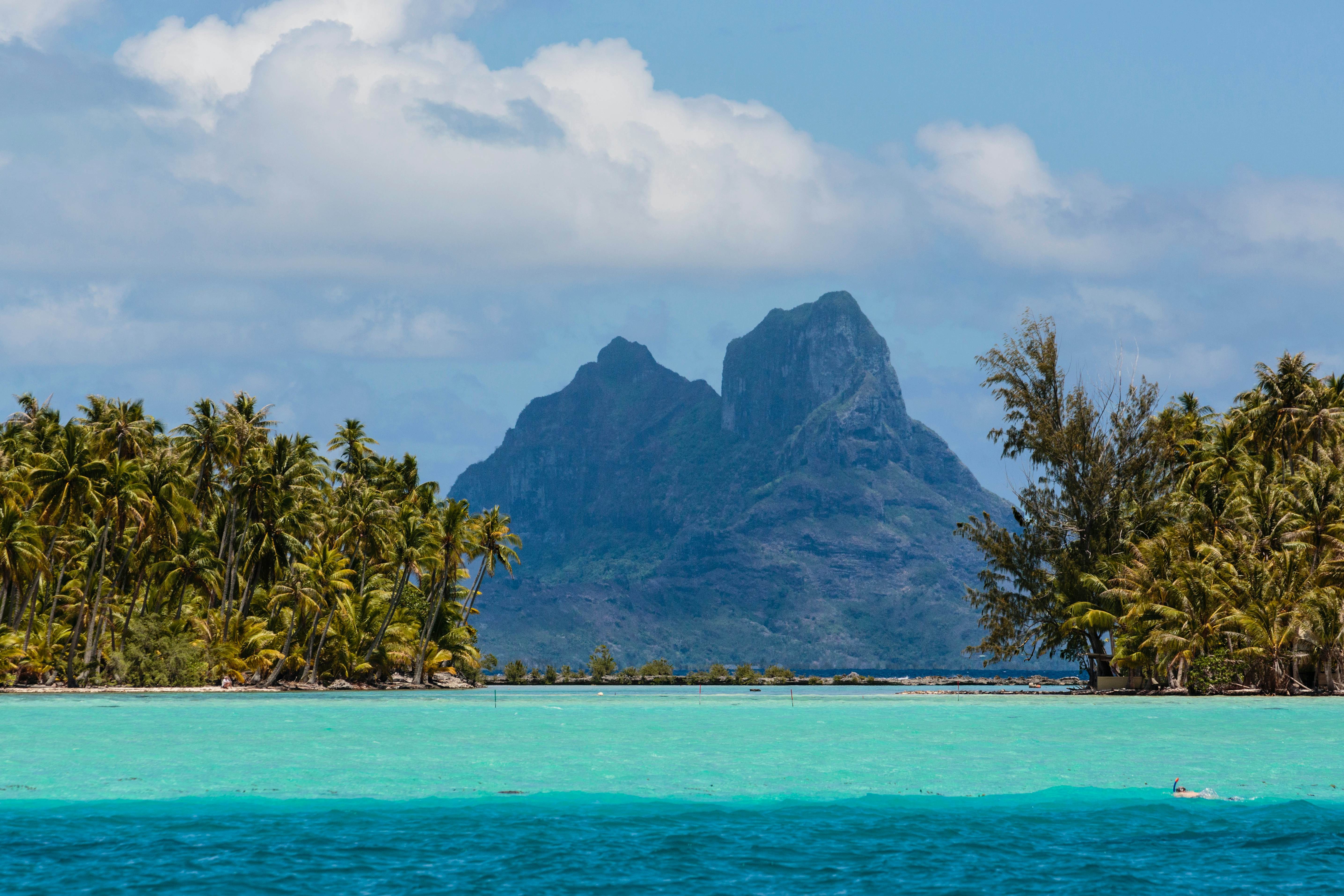 Two mountains centered behind a turquoise and blue lagoon, and flanked by green palm trees.