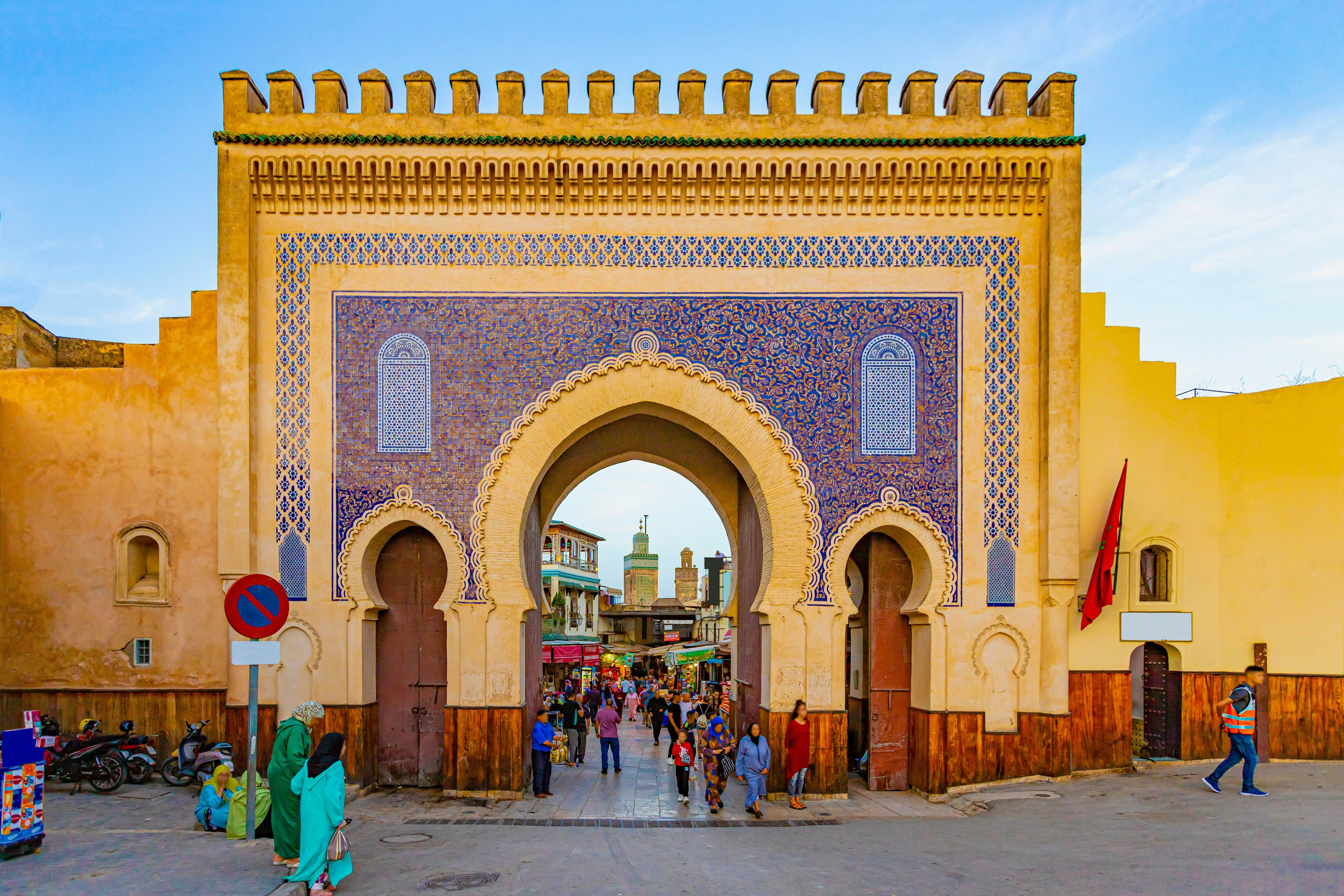 A gate with three rounded entryways, with people walking through the largest one in the center to the street on the other side; the tan stone is decorated with geometric shapes using tiles in shades of blue.