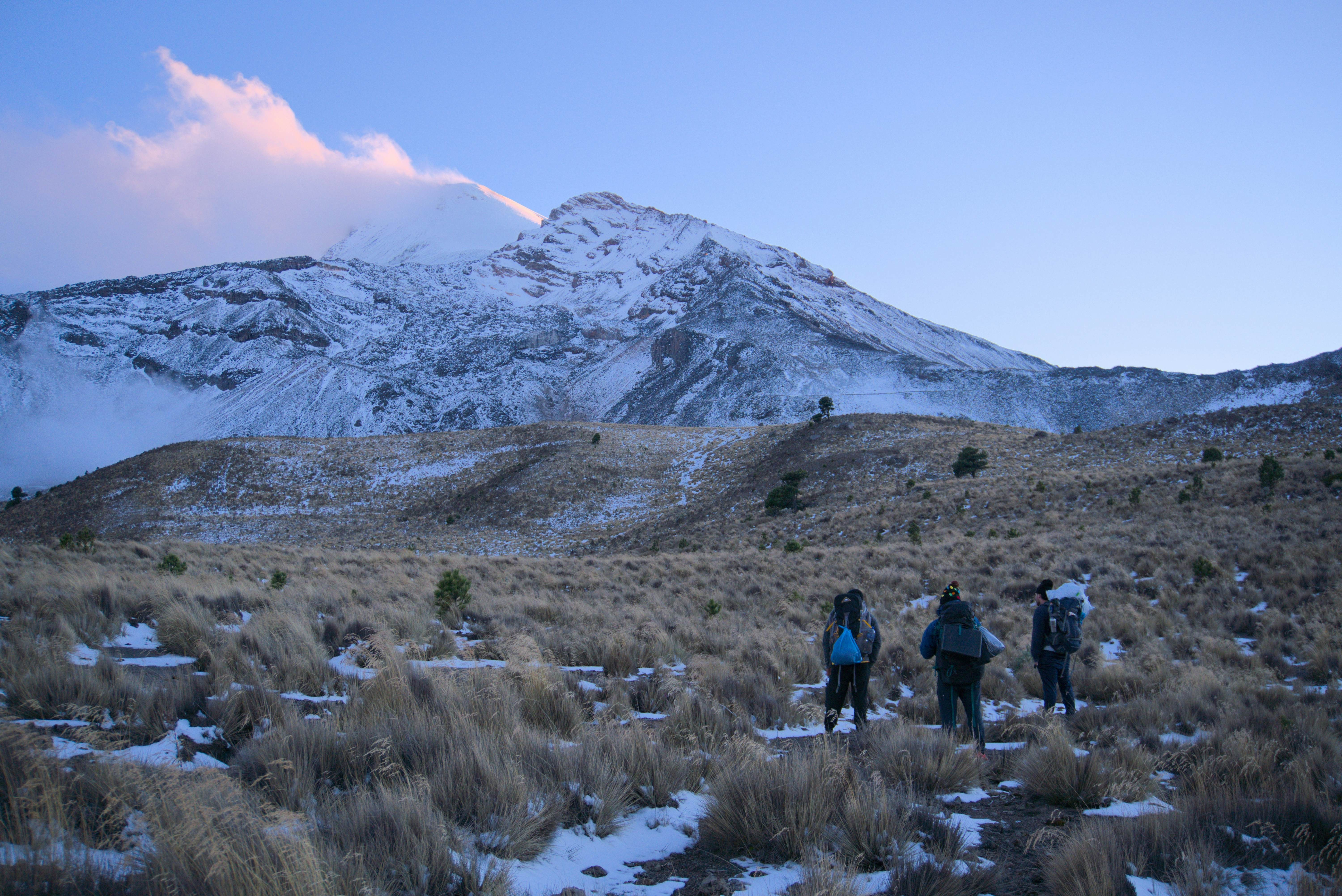 People hike across a snow-dusted field toward the snowy peak of a mountain at dawn.