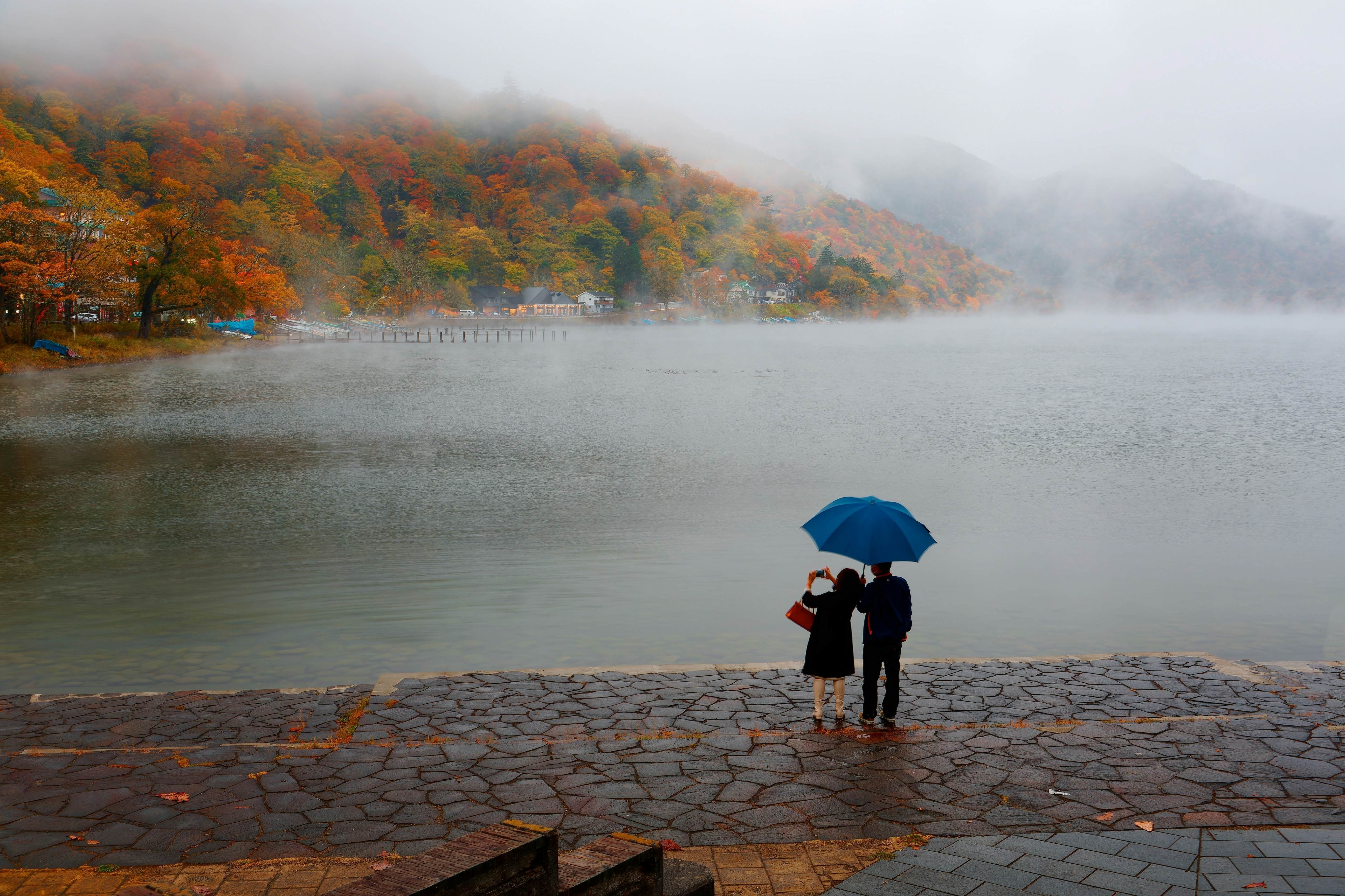 Two people stand under an umbrella on the shore of a lake: Overcast weather at Lake Chuzenji