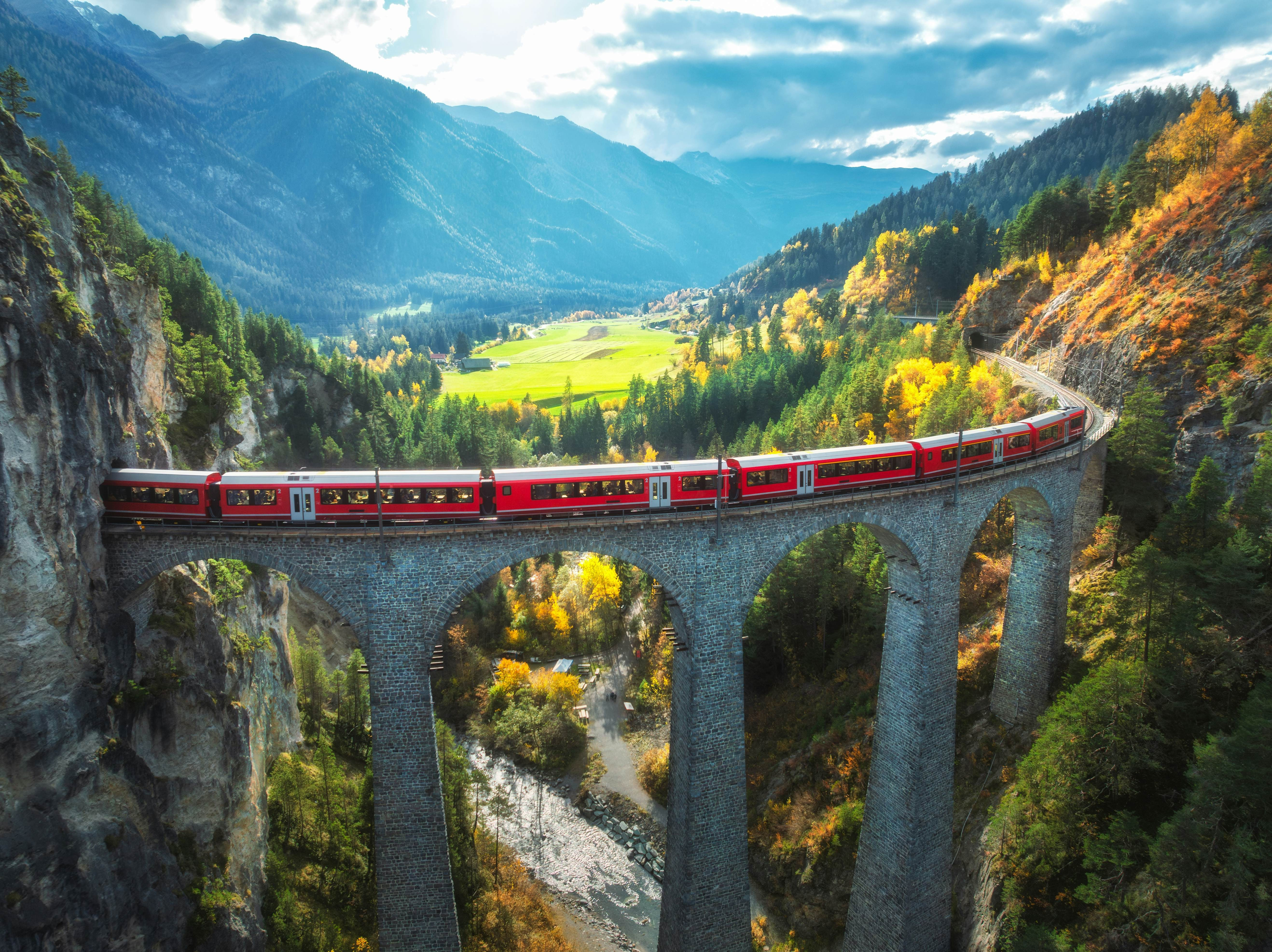 Red train on a viaduct going into mountain tunnel with autumnal forest and mountains in the distance and sun shining through clouds.