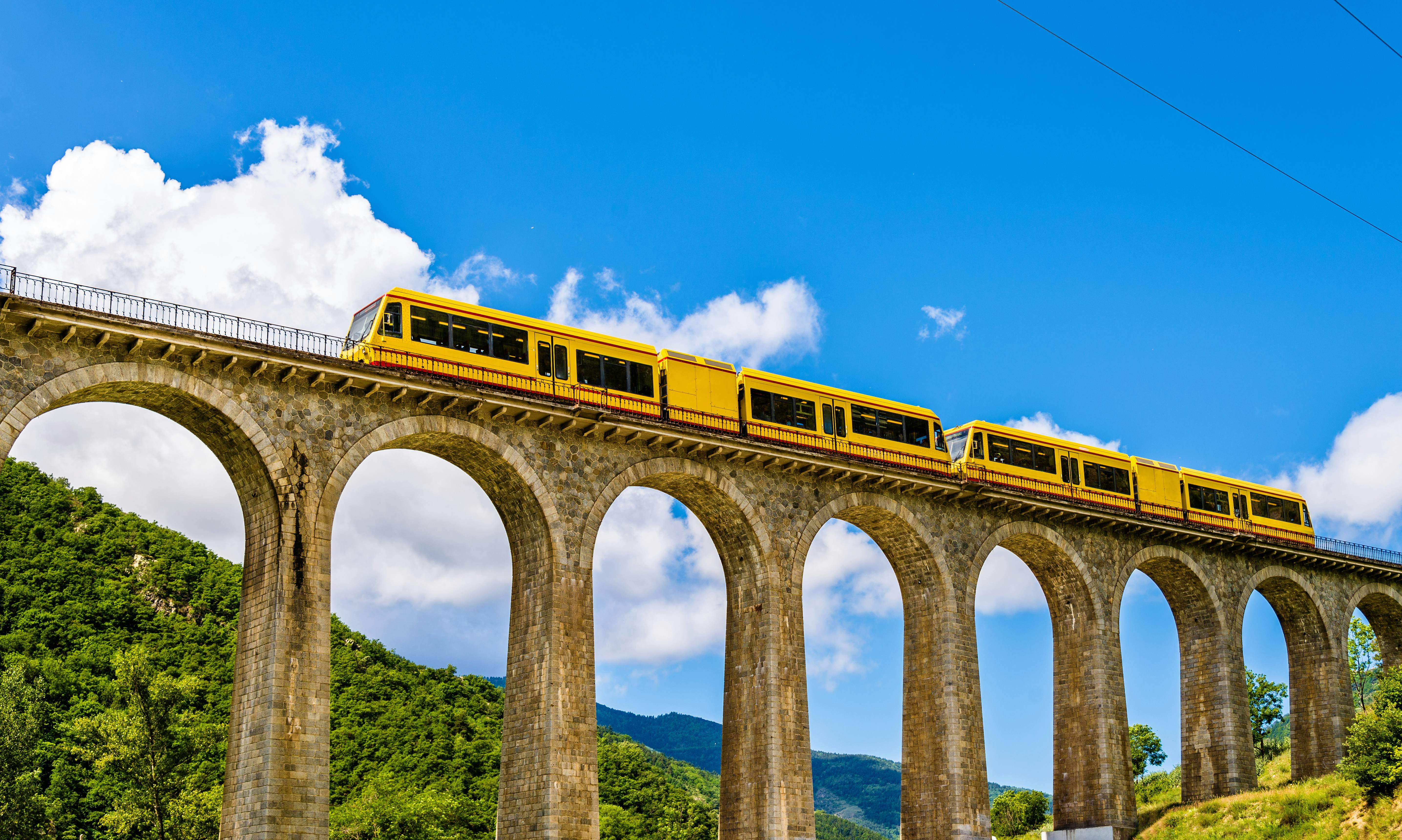 Yellow train on a raised ston bridget with green trees below and mountains in the distance on a sunny day.