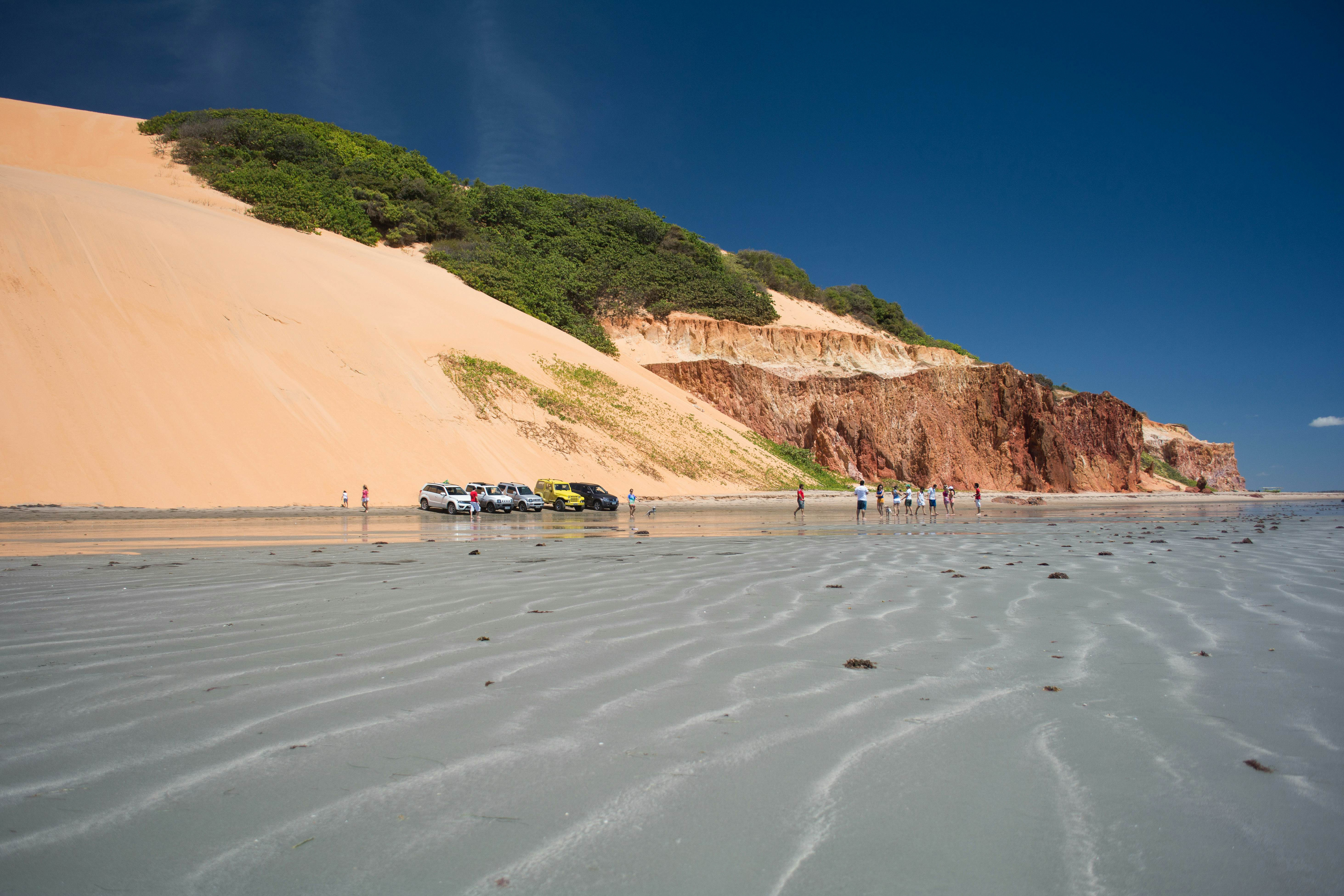 A wide view of shallow waves lapping a beach, with people and vehicles parks. Large sand dunes and cliffs rise from the beach.