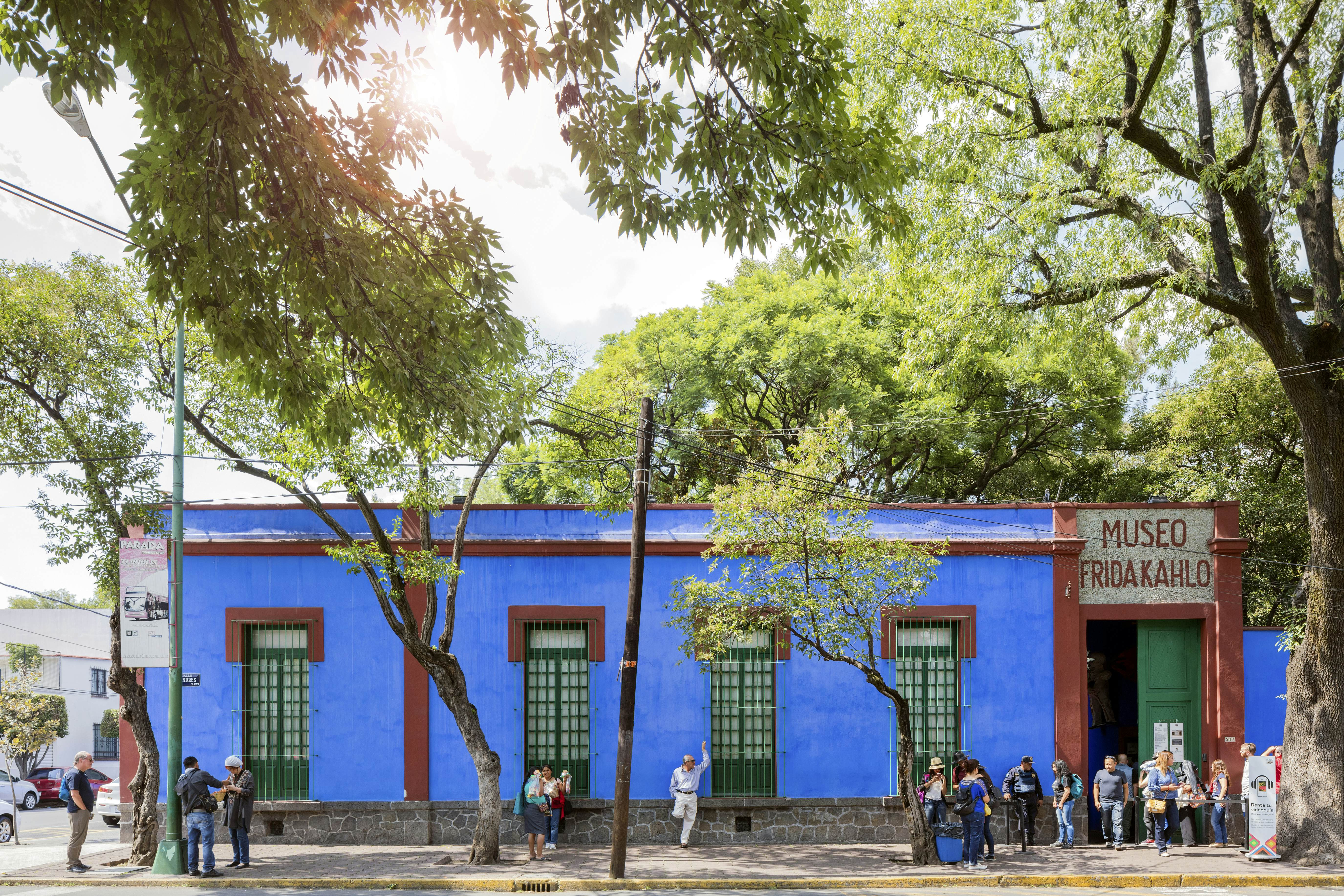 People line up in front of a one-story building painted blue on a city street shaded by trees