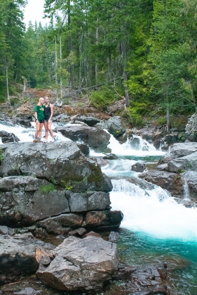 two teen girls standing on a rock with glacier aqua colored waterfalls below them and green forests behind them
