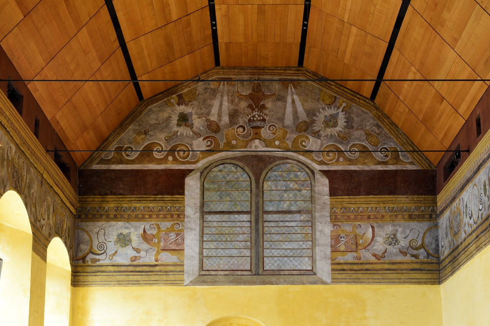 The upper part of a tall room, with large, arched windows. The top of th walls are painted with a mural featuring windows, waves and other designs. The ceiling is arched and clad in wood. The ceiling is arched and clad in wood.
