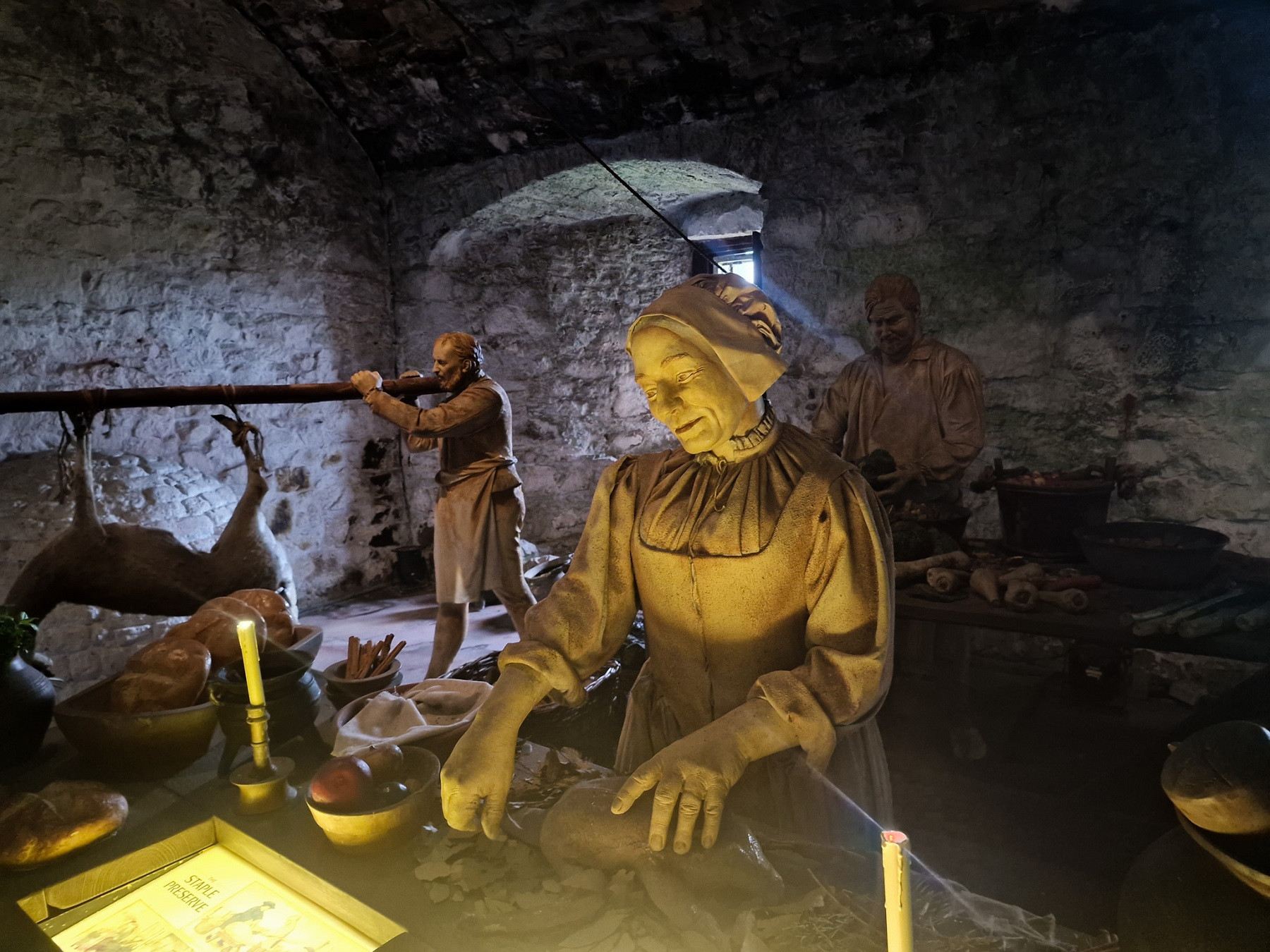 Tables in the stone kitchen, with apples, bread and other foodstuffs on them. Wax models stand behind the tables, preparing food. One model is roasting a deer on a spit. 