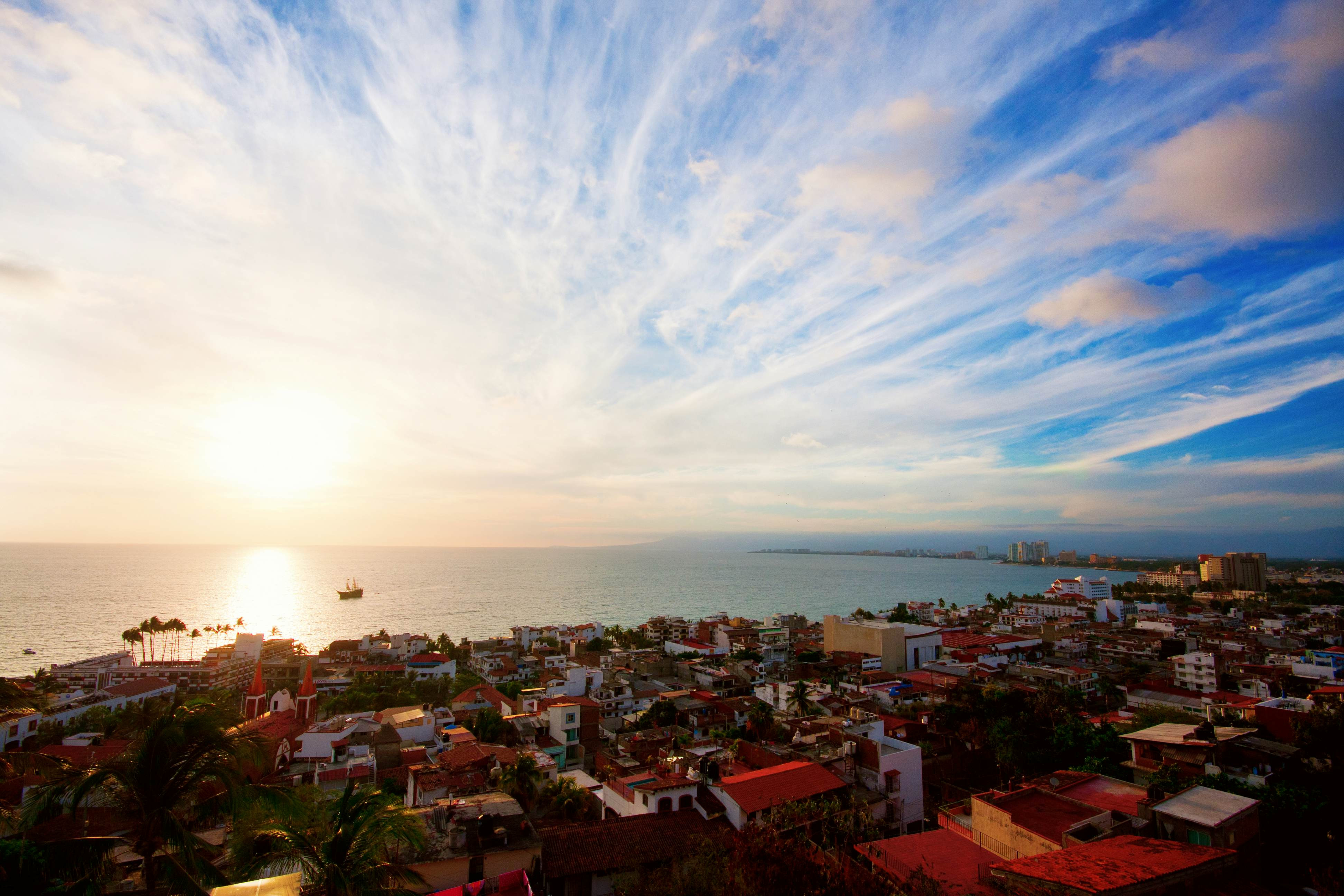 A high-angle view of the sun setting over a bay, shining light on the roofs of a seaside city. A double-masted boat is moored in the bay.