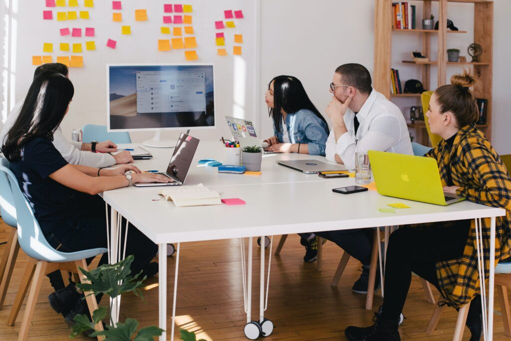 A team of 5 people sit at tables in a n office, looking at a monitor and post-it notes on the wall.