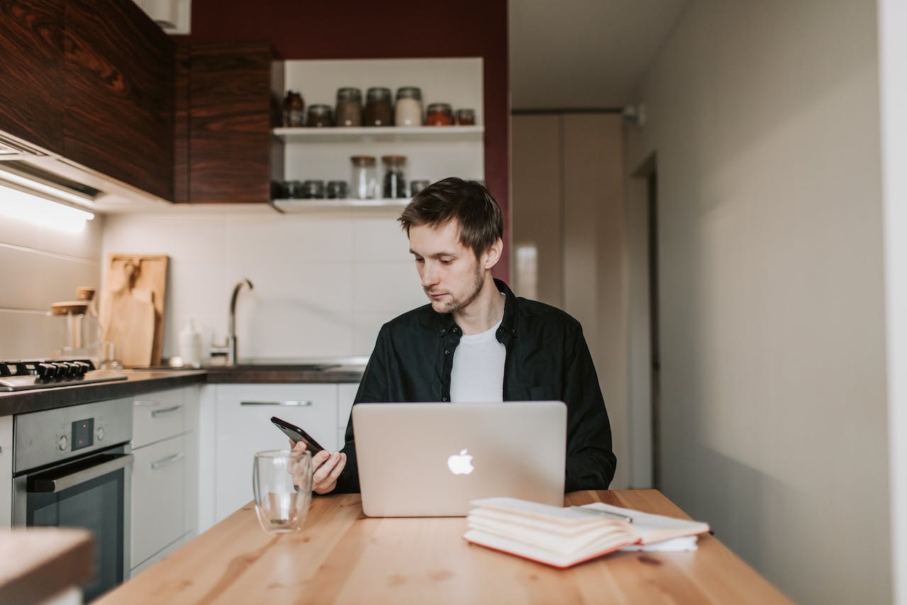 thoughtful-male-freelancer-using-smartphone-and-laptop-in-kitchen