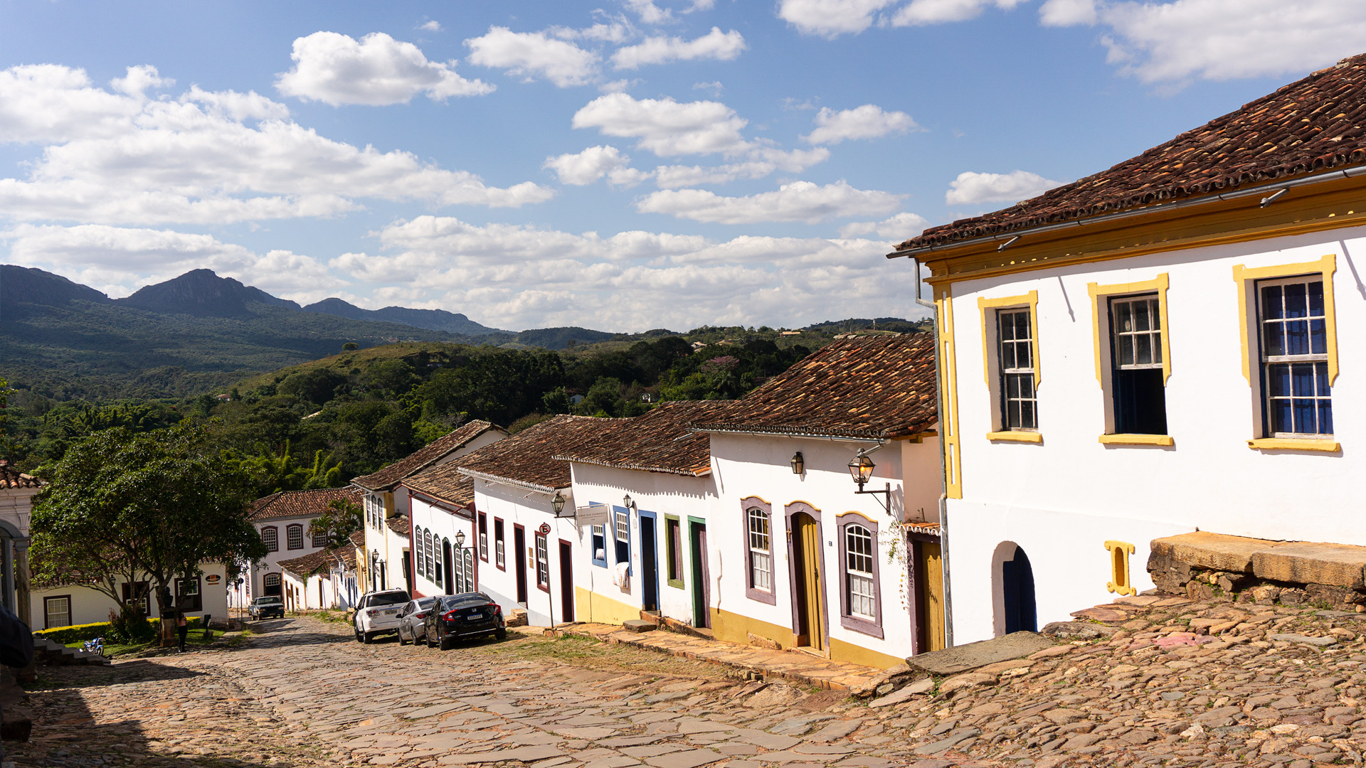 A cobblestone street going downhill, lined with colonial style houses (white with yellow trim), and tiled roofs. tiradentes, brazil.