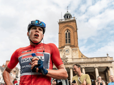 Paul Magnier, winner of Stage 5 of the Tour of Britain Men 2024, wearing a red cycling jersey, stands in front of All Saints Church following the race
