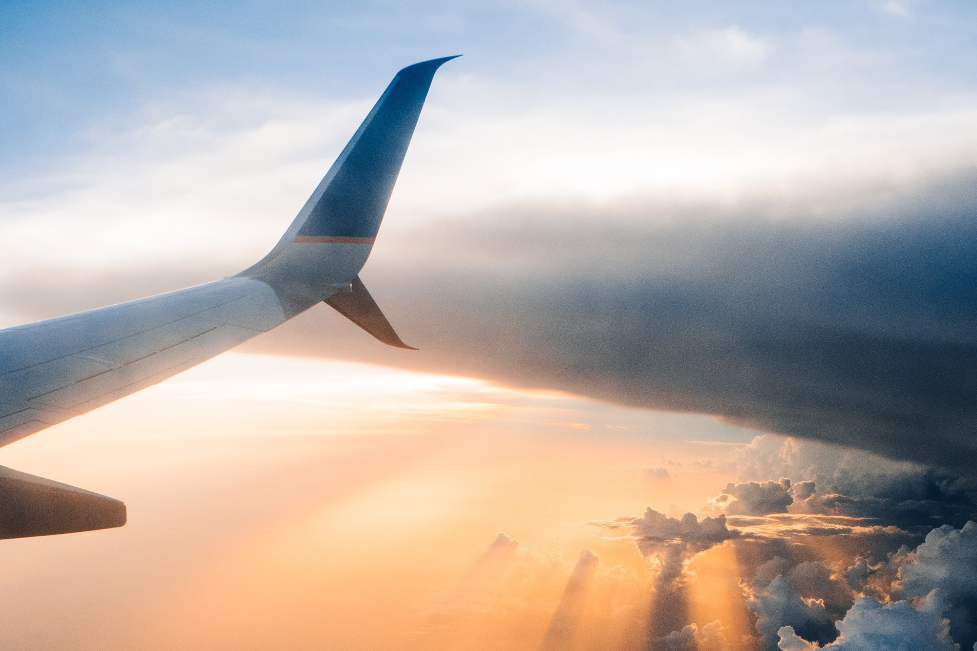 The wing of a passenger jet is visible from the view of an airline seat with brilliant sunshine breaking through dark clouds