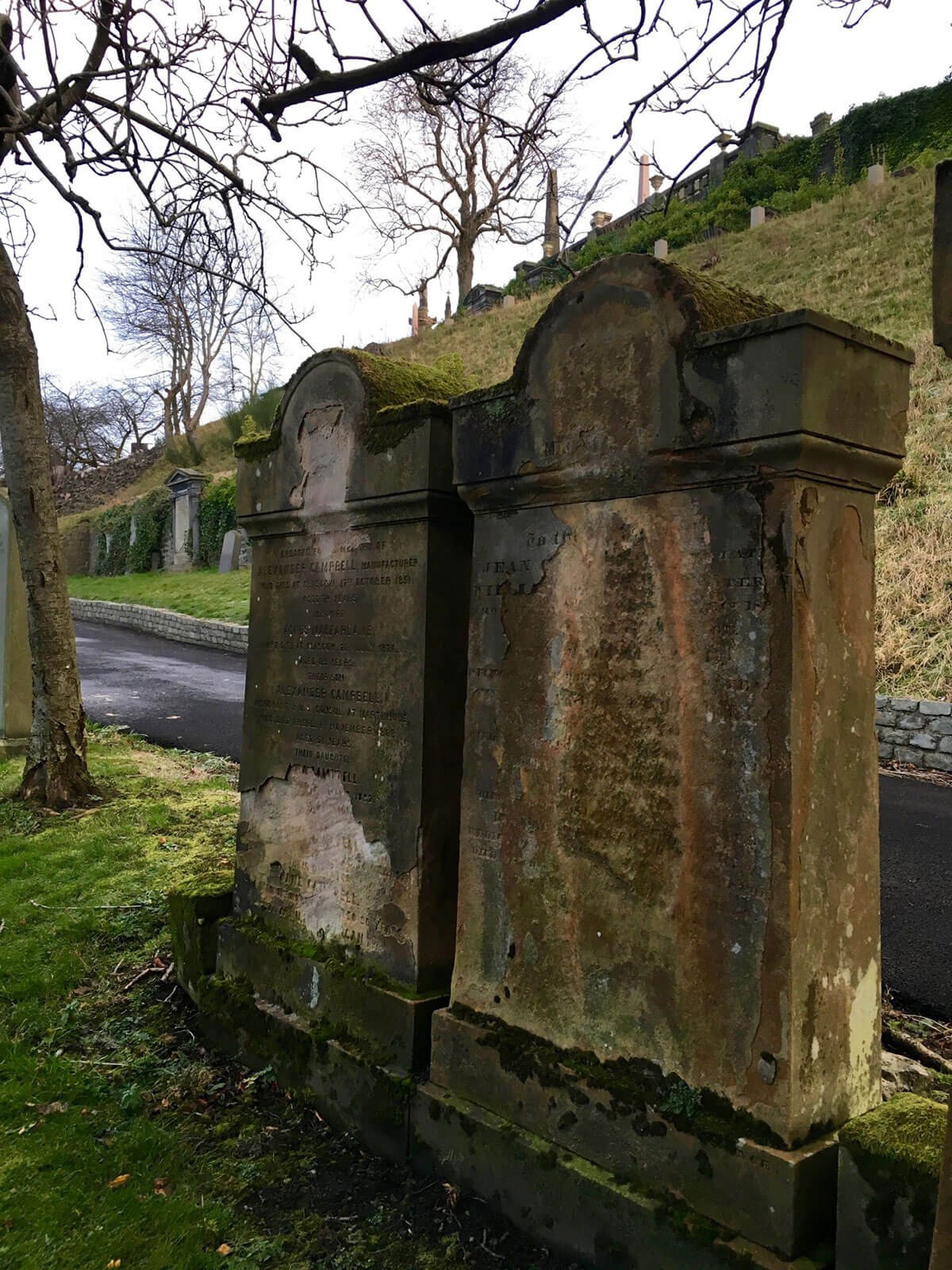 Two weathered, mossy graves stand side-by-side in Glasgow Necropolis, their inscriptions faded with time, under an overcast sky, evoking a sense of history and tranquility.