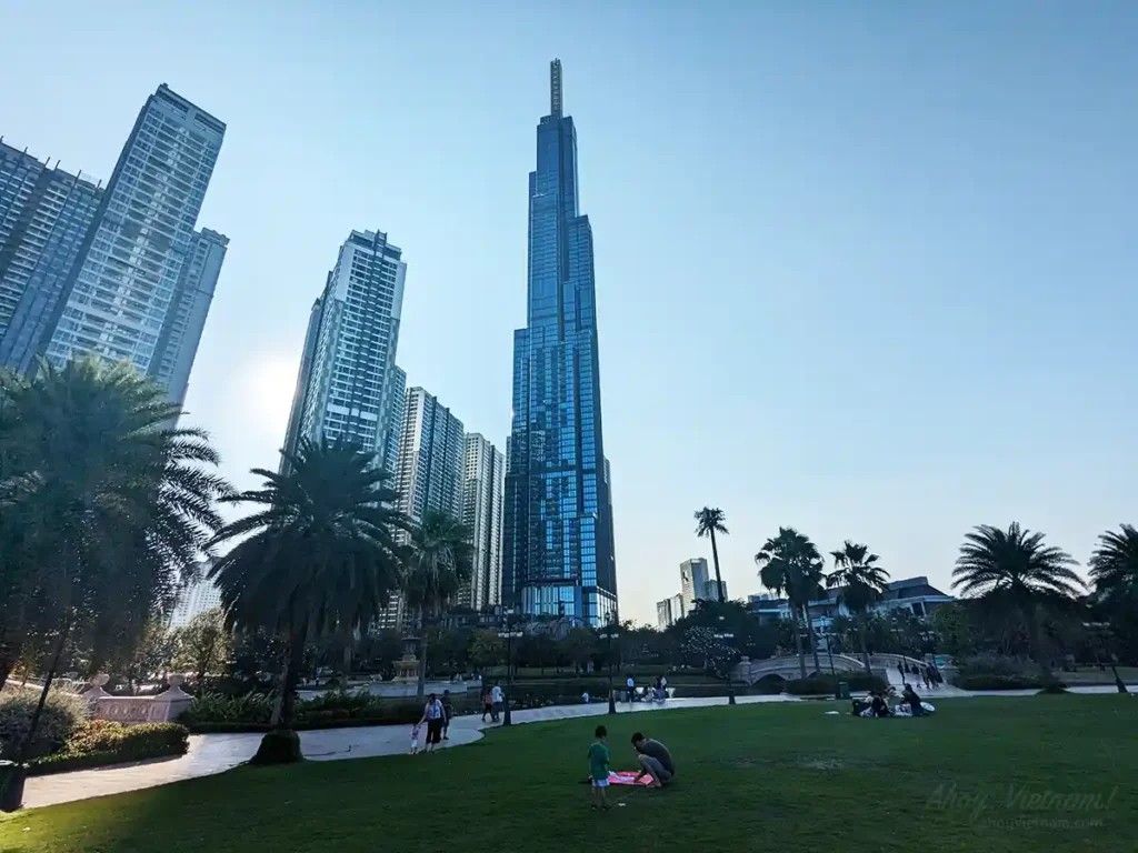 Vinhomes Central Park with grass, families, apartments on the left and the Vinpearl Landmark 81 building in the distance.