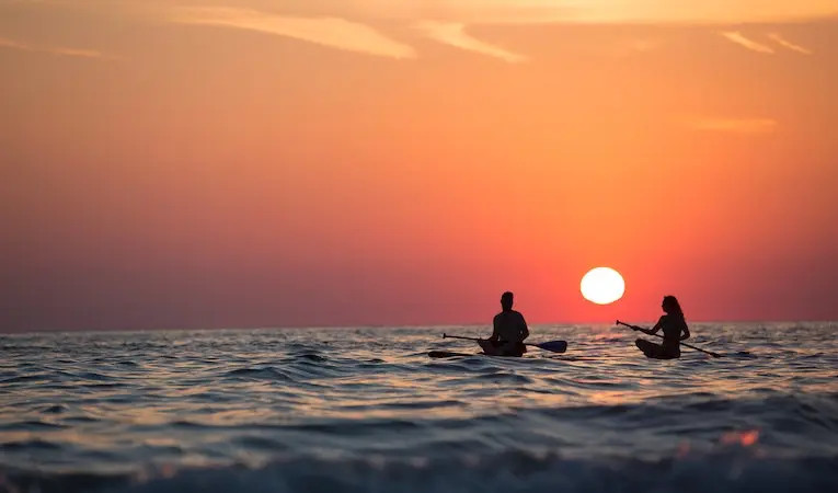 Two people paddleboarding on a calm ocean during sunset, highlighting the adventurous lifestyle