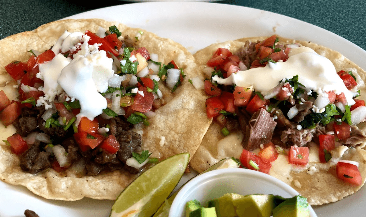 two carne asada tacos with two slices of lime and a side bowl of diced avocado