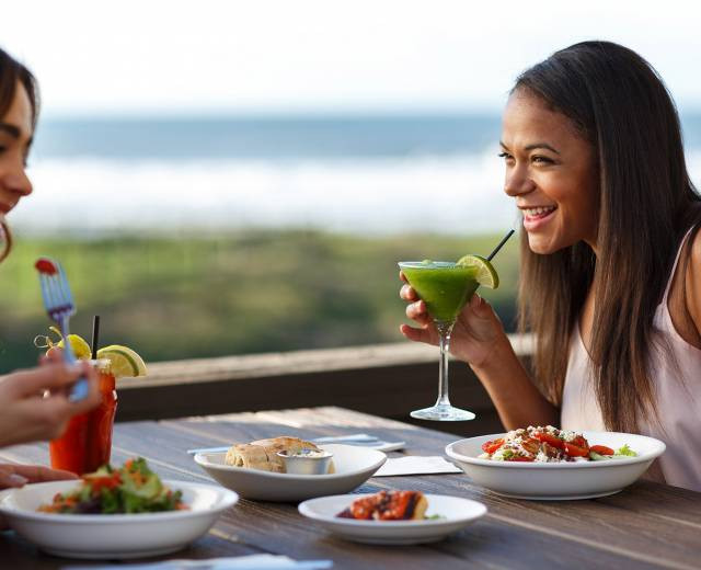 Two women enjoy food and drinks at a table outdoors at a Lincoln City restaurant, smiling and laughing